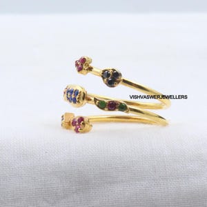 Pu&ograve; includere: Un anello a spirale dorato con accenti di pietre preziose colorate. L'anello presenta piccoli gruppi di pietre rosse, blu e nere e un motivo a foglia verde. Il testo "VISHVASWERJEWELLERS" &egrave; visibile.