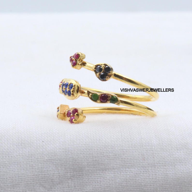 Pu&ograve; includere: Un anello a spirale dorato con accenti di pietre preziose colorate. L'anello presenta piccoli gruppi di pietre rosse, blu e nere e un motivo a foglia verde. Il testo "VISHVASWERJEWELLERS" &egrave; visibile.
