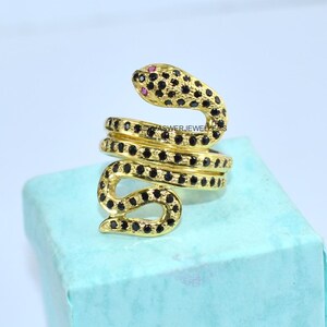 May include: A gold-toned snake ring with black and red accents. The snake's body coils around the finger, with black stones covering the body and a red stone for the eye. The ring is on a light blue surface.