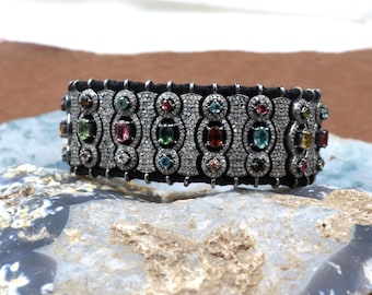 May include: A black leather bracelet with a silver and diamond design. The bracelet is adorned with colorful gemstones, including pink, green, and blue.