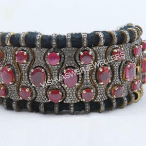 May include: A silver and gold bracelet with a black cord and red gemstones. The bracelet is decorated with small diamonds and features the text "VISHVASWER JEWELLERS".