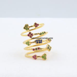May include: A gold-colored spiral ring with multiple levels, each adorned with colorful gemstones. The gemstones are arranged in floral and geometric patterns, including green, red, and blue stones. The ring is set against a white background.