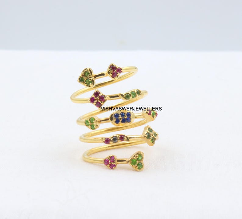 May include: A gold-colored spiral ring with multiple levels, each adorned with colorful gemstones. The gemstones are arranged in floral and geometric patterns, including green, red, and blue stones. The ring is set against a white background.