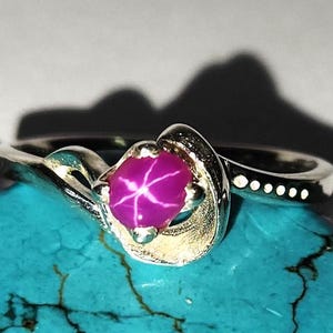 Può includere: Un anello d'argento con una pietra preziosa di rubino stellato rosa incastonata in un design a spirale. L'anello è su uno sfondo turchese.