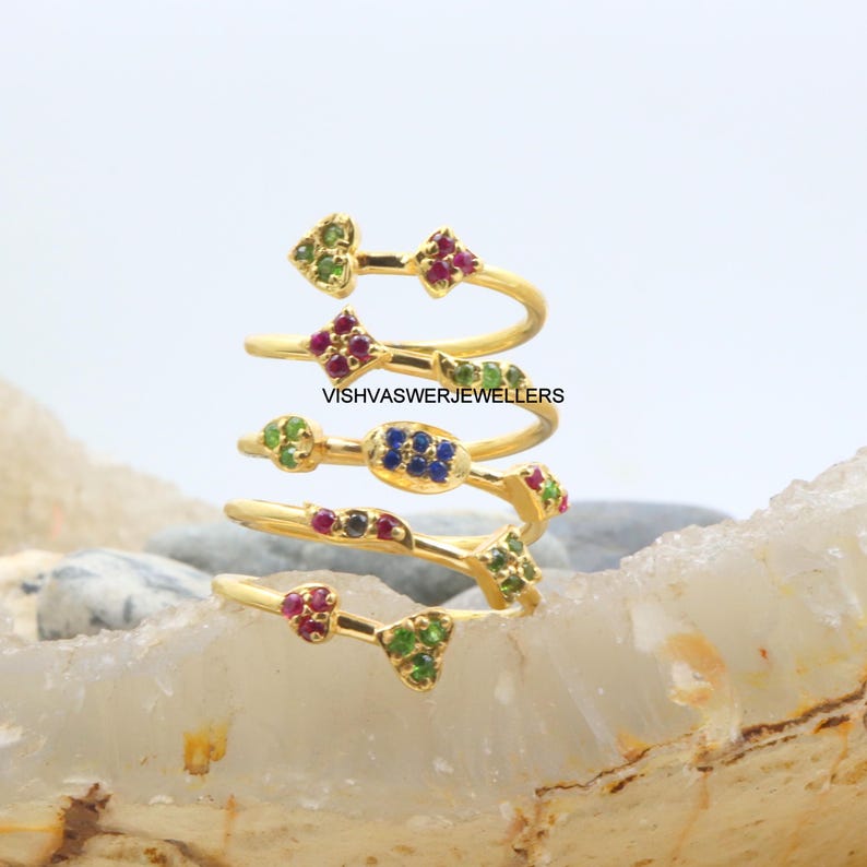 May include: A gold-colored spiral ring with multiple bands, each adorned with colorful gemstones. The gemstones are arranged in clusters of red, green, and blue. The text "VISHVASWERJEWELLERS" is visible on the ring.