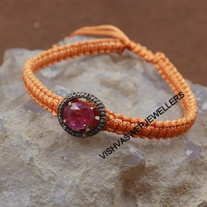 May include: A pink gemstone set in a silver bezel with a diamond halo, on a braided orange cord bracelet. The bracelet is tied with a knot and the text "VISHVAS JEWELLERS" is visible.