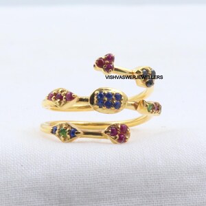 Pu&ograve; includere: Un anello color oro con un design a spirale, adornato con piccole pietre preziose colorate. L'anello presenta pietre rosse, blu e verdi e il testo "VISHVASWERJEWELLERS". L'anello &egrave; posto su uno sfondo bianco.