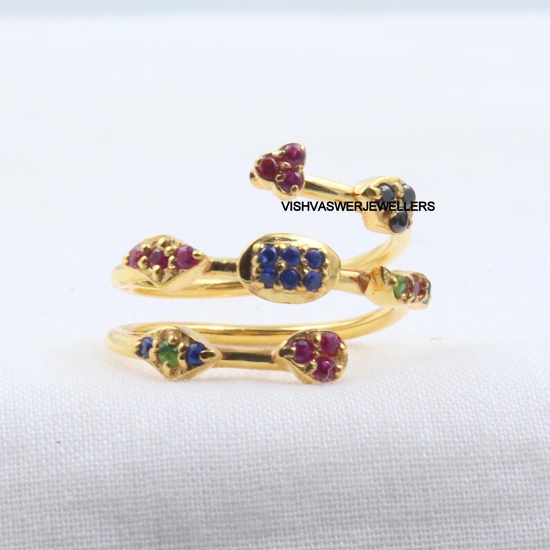 Pu&ograve; includere: Un anello color oro con un design a spirale, adornato con piccole pietre preziose colorate. L'anello presenta pietre rosse, blu e verdi e il testo "VISHVASWERJEWELLERS". L'anello &egrave; posto su uno sfondo bianco.