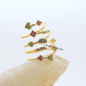 May include: A gold-colored spiral ring with multiple levels, each adorned with colorful gemstones. The gemstones are arranged in various shapes, including hearts, squares, and clusters, in shades of red, green, and blue. The ring is displayed on a light-colored, textured surface.