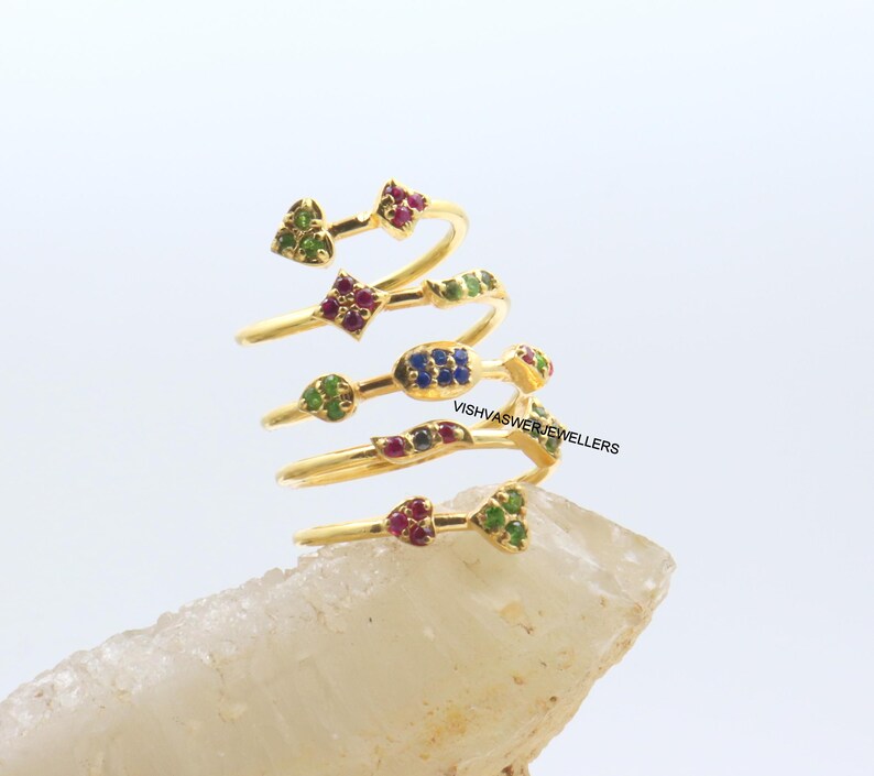 May include: A gold-colored spiral ring with multiple levels, each adorned with colorful gemstones. The gemstones are arranged in various shapes, including hearts, squares, and clusters, in shades of red, green, and blue. The ring is displayed on a light-colored, textured surface.