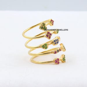 May include: A gold-colored spiral ring with colorful gemstone accents. The ring features a coiled design with small clusters of green, red, and blue gemstones. The text "VISHVASWERJEWELLERS" is visible.
