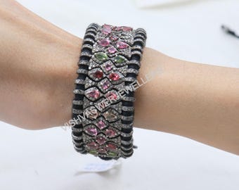 May include: A wide, ornate bracelet featuring a black band with rows of colorful gemstones. The gemstones are a mix of pink, green, and red, set in a silver-toned metal. The bracelet is approximately 2 inches wide.