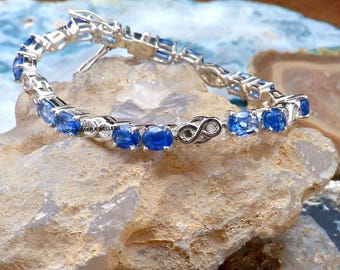 May include: A silver bracelet with oval-cut, vibrant blue gemstones. The bracelet is set on a light-colored stone surface. The clasp features a decorative infinity symbol. The text "VISHWAS JEWELLERS" is visible on the bracelet.