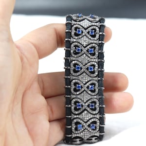 Puede incluir: Un brazalete de plata con una banda negra y un diseño de corazones entrelazados y símbolos de infinito. El brazalete está adornado con pequeños diamantes transparentes y zafiros azules.
