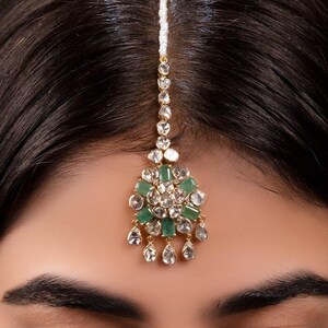 May include: A gold and silver forehead ornament with green and clear stones. The ornament is worn on the forehead and has a long chain that hangs down the back of the head.
