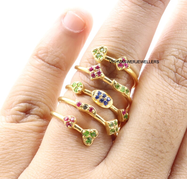 May include: Gold rings with colorful gemstones, including green, red, and blue stones. The rings have unique designs, with some featuring heart and square shapes. The rings are displayed on a finger, showcasing the jewelry's intricate details and craftsmanship.