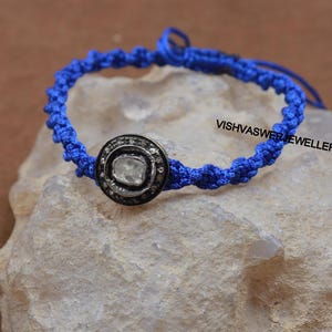 May include: A blue braided cord bracelet with a silver and white diamond accent. The diamond is set in a round bezel setting and surrounded by smaller diamonds. The bracelet is tied with a knot and the text "VISHVASWER JEWELLERS" is visible in the background.