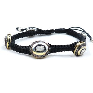 May include: A black braided bracelet with three oval-shaped charms. Each charm is set with a large, clear diamond surrounded by smaller diamonds. The charms are gold-toned and have a silver-toned border.