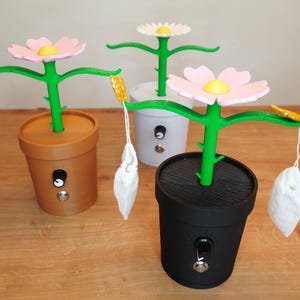 Op de afbeelding: Drie bloemvormige thee-infusers in bruine, grijze en zwarte potten. Elke bloem heeft een roze of wit bloembladontwerp met een geel centrum en een groene stengel. Aan elke arm van de bloem hangt een theezakje.