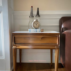 May include: A vintage wooden end table with a single drawer and tapered legs. The table top is white and the drawer is brown. A clock and two glass bottles are on top of the table.