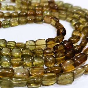 Puede incluir: Hebras de cuentas de piedras preciosas verde oliva y marrón. Las cuentas son rectangulares y redondeadas, con un aspecto pulido y translúcido. Las cuentas están ensartadas, creando un efecto en capas. La paleta de colores incluye tonos de verde, marrón y ámbar.