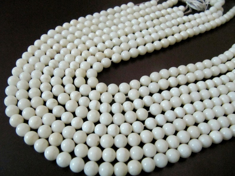 May include: A strand of white round beads, made of natural shell, with a smooth, polished finish. The beads are strung on a clear thread and are arranged in a circular pattern.
