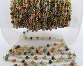 May include: A spool of gold chain with small, faceted gemstone beads in various colours, including red, green, blue, and purple. The chain is laid out in a straight line, showing the beads and chain links.