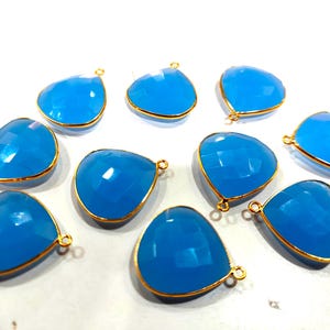 May include: Ten teal blue gemstone pendants with gold-toned metal settings. Each pendant is shaped like a teardrop and has a small loop at the top for attaching to jewelry.