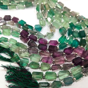 May include: Strands of colorful fluorite beads in shades of green, purple, and clear. The beads are rectangular and strung together with silver-colored metal beads. Green tassels are visible at the bottom of the image.
