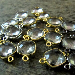 Puede incluir: Una colección de conectores de piedra preciosa de cristal transparente con bucles de metal en tonos dorados y plateados. Los conectores son de forma cuadrada y tienen un diseño facetado.