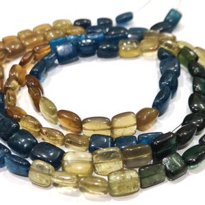 Puede incluir: Cadenas de cuentas de piedras preciosas en tonos azules, verdes y ámbar. Las cuentas, de formas rectangulares y ovaladas, están ensartadas para formar collares. Las cuentas pulidas reflejan la luz.