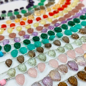 Könnte beinhalten: Eine Strähne aus Edelsteinperlen in verschiedenen Farben, darunter Amethyst, Rosenquarz, grüner Aventurin und Rauchquarz. Die Perlen sind tropfenförmig und auf einem Silberdraht aufgefädelt. Jede Perle hat ein geschnitztes Blattdesign.