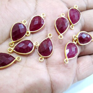 May include: Ten gold-plated ruby gemstone connector charms. Each charm features a teardrop-shaped ruby gemstone with a faceted finish. The charms have two jump rings for attaching to jewelry.