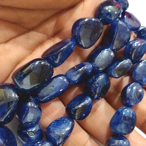 May include: A strand of dark blue gemstone beads, each bead is a smooth, oval shape. The beads are strung together on a clear thread and are displayed on a hand.