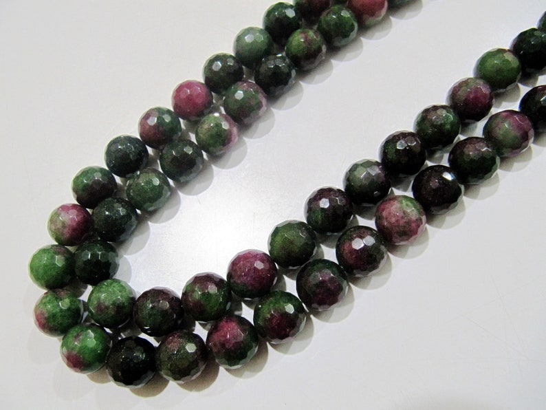 May include: A strand of green and pink gemstone beads. The beads are faceted and have a round shape. The beads are strung on a clear thread.