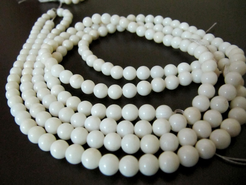 May include: A strand of white shell beads, strung on a white thread. The beads are round and smooth, and they are arranged in a circular pattern.