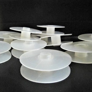 May include: A stack of white plastic spools for thread or yarn. The spools are stacked in a random pattern on a black surface.