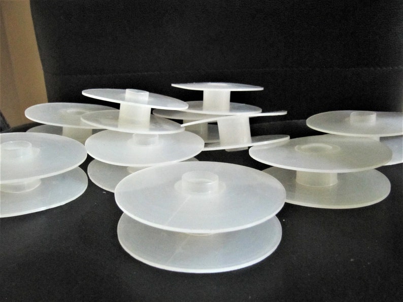 May include: A stack of white plastic spools for thread or yarn. The spools are stacked in a random pattern on a black surface.