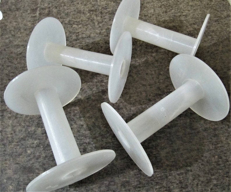 May include: Four white plastic spools for thread or yarn. The spools are all the same size and shape, with a large, flat circular base and a smaller, cylindrical center.
