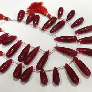 Op de afbeelding: Slingers van dieprode, druppelvormige kralen, geregen op zilverkleurig draad. De kralen hebben een glanzende afwerking en zijn in een cirkelvormig patroon gerangschikt. Rode kwasten zijn aan de uiteinden bevestigd. Geschikt voor het maken van sieraden.