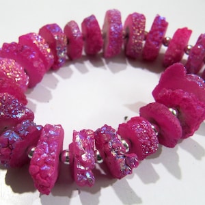 Puede incluir: Un brazalete de cristal rosa con acentos iridiscentes y un borla de plata. El brazalete está hecho de cuentas de cristal natural y está enhebrado en una cadena de plata.