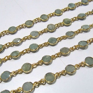 Puede incluir: Una hebra de cadena de oro con piedras ovaladas de color azul claro. La cadena est&aacute; dispuesta en cuatro filas sobre una superficie blanca.