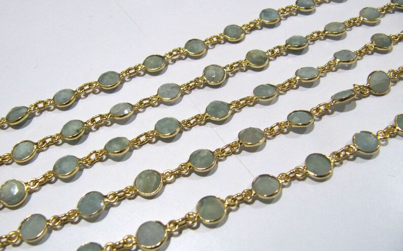 Puede incluir: Una hebra de cadena de oro con piedras ovaladas de color azul claro. La cadena est&aacute; dispuesta en cuatro filas sobre una superficie blanca.