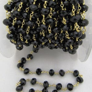 May include: Black faceted glass beads strung on a gold chain. The beads are small and round, and the chain is delicate. This is a strand of beads for jewelry making.
