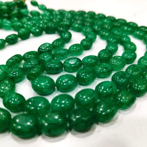 Peut inclure: Un collier de perles de pierres précieuses de couleur vert émeraude de forme ovale. Les perles sont lisses et polies, et elles sont enfilées sur un fil transparent.