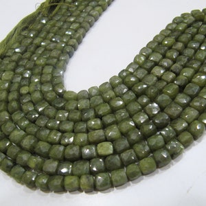 以下が含まれることがあります： オリーブグリーンの宝石ビーズのクローズアップ。各ビーズはファセットキューブで、キラキラとした効果を生み出しています。ビーズは一緒にひもでつながれており、ネックレスまたはブレスレットを形成しています。色は落ち着いたアースカラーのグリーンです。