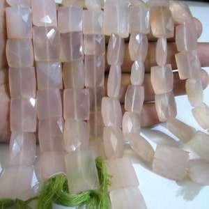 May include: A strand of pale pink faceted gemstone beads. The beads are square shaped and have a translucent appearance. The beads are strung on a cord with green tassels at the ends.