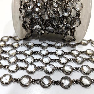 Puede incluir: Una cadena de plata con cuentas de cristal facetadas transparentes. La cadena está enrollada y dispuesta en filas, mostrando el detalle de la cadena y las cuentas.