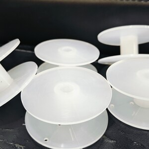 May include: Five white plastic bobbins for thread spools. The bobbins have a center hole and a raised lip around the edge.