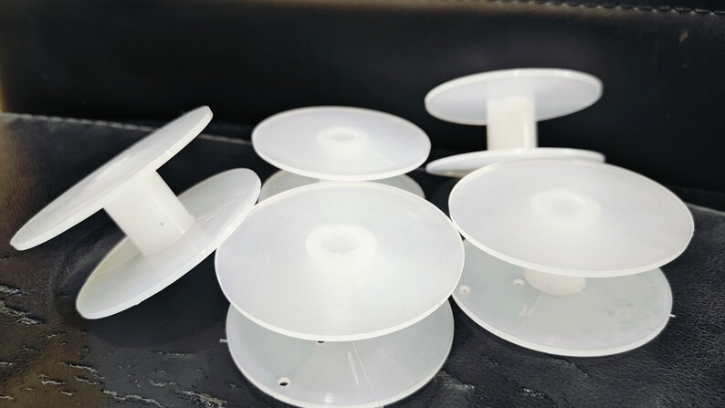 May include: Five white plastic bobbins for thread spools. The bobbins have a center hole and a raised lip around the edge.